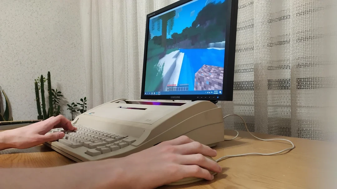 Un joueur utilise un PC conçu à partir d’une ancienne machine à écrire pour jouer à Minecraft, avec un écran affichant une scène en bord de rivière.