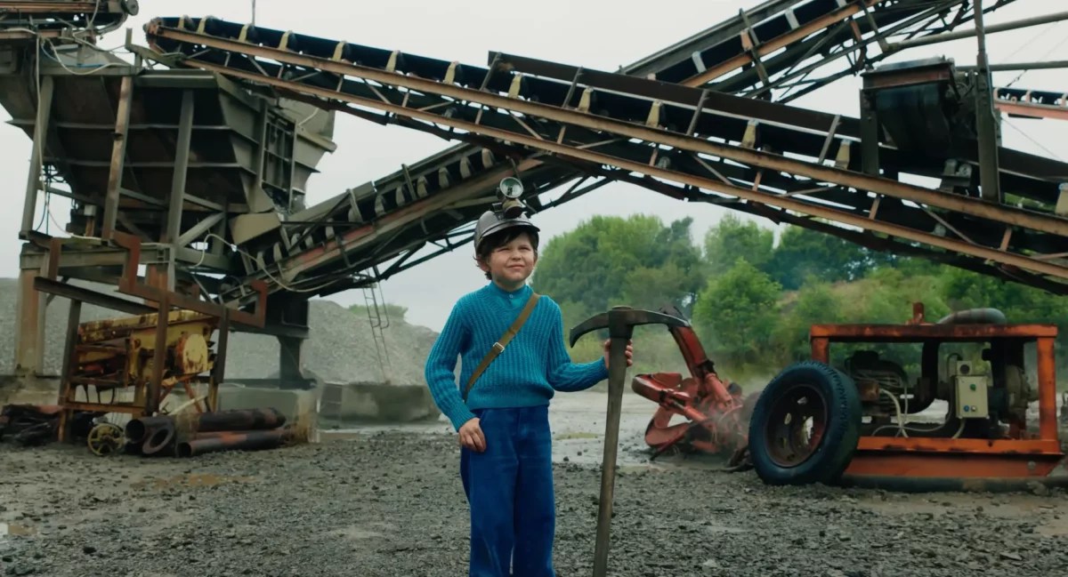 Jeune Steve dans le film Minecraft, portant un pull bleu et tenant une pioche dans une carrière, évoquant ses débuts de mineur sur Terre.